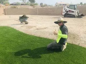 In this image from Infinity Concepts Landscaping in Bullhead City, AZ, a person kneels on a patch of artificial turf, giving a thumbs up. Behind them, a wheelbarrow and compact loader are visible along with a brick wall, highlighting thoughtful backyard design and unique landscaping elements. This setup underscores the focus on professional landscape design services provided by Infinity Concepts Landscaping.